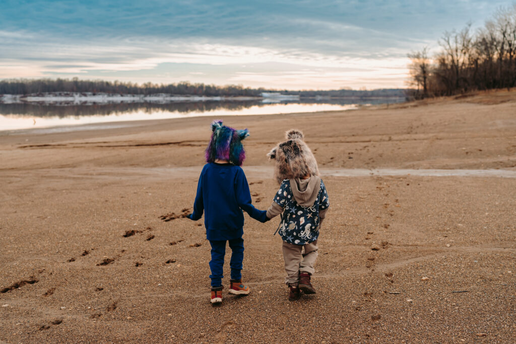 Two little boys walk down a beach holding hands away from the camera. Both are wearing furry wolf hats
