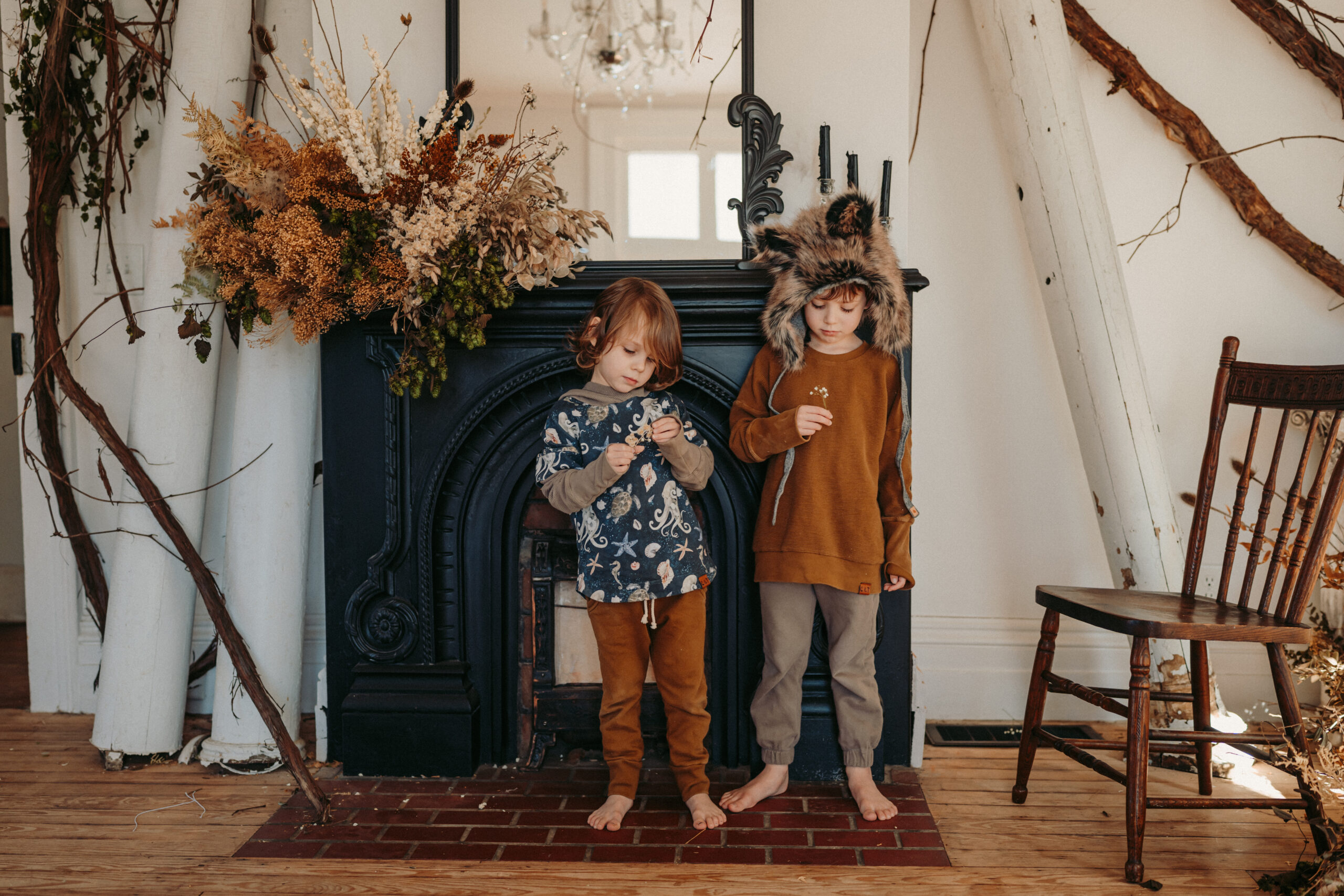 Two boys wearing furry animal bonnets stand in front of a black fireplace surrounded by dried flowers.
