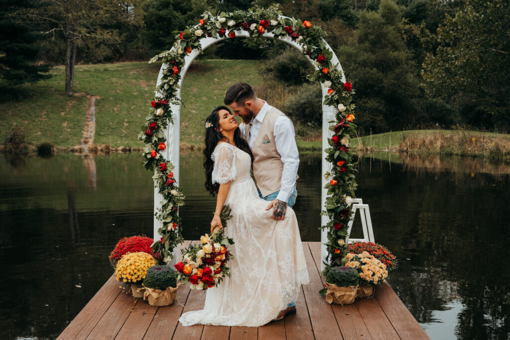 Bride and groom stand in front of archway covered in flowers after getting married. They're on a dock to a pond and are surrounded by flowers, and looking lovingly at each other.